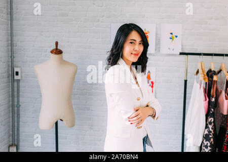 Contenuto Asian fashion designer permanente al tavolo e avente bracci incrociati con attrezzatura per cucire e sorridente alla fotocamera in officina di luce Foto Stock