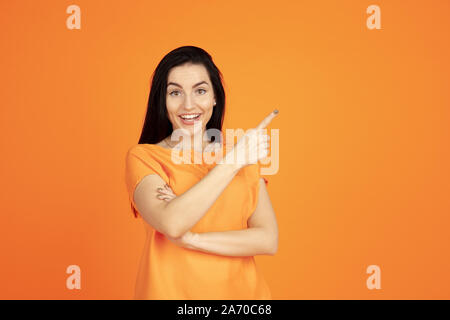 Giovani caucasici ritratto di donna su arancione di sfondo per studio. Bellissima femmina bruna modello in camicia. Concetto di emozioni umane, l'espressione del viso, vendite, annuncio. Copyspace. Dispositivo di puntamento, mostrando un sorriso. Foto Stock