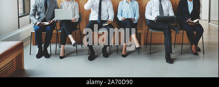 Diversi gruppi di imprenditori irriconoscibile seduto in una fila di sedie nella zona della reception di un ufficio in attesa che il loro colloquio gli appuntamenti Foto Stock