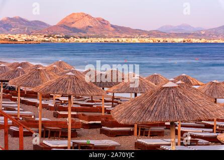 I tetti di paglia di ombrelloni in legno e sedie a sdraio con materassi su una spiaggia deserta del lungomare di raggi del tramonto. Foto Stock