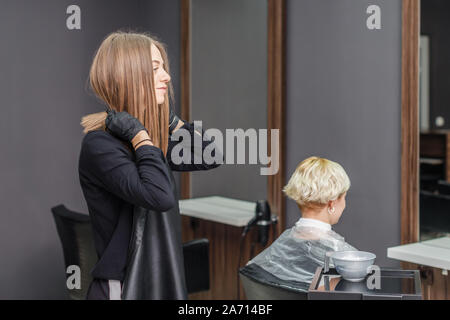 La femmina professionale parrucchiere mette sul grembiule nero per tingere la donna capelli nel salone di parrucchiere. Foto Stock