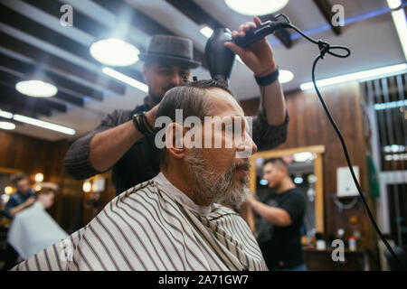 Il nonno si ottiene un taglio di capelli al parrucchiere in un barbiere. Taglio di capelli alla moda di un uomo vecchio, titolare di una pensione o di una rendita nel negozio del barbiere. Capelli e barba care. Russia, S Foto Stock