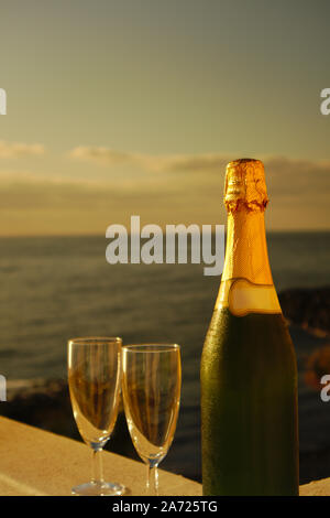 Una bottiglia di champagne e due bicchieri per qualche celebrazione al tramonto sul mare che è visto in background Foto Stock
