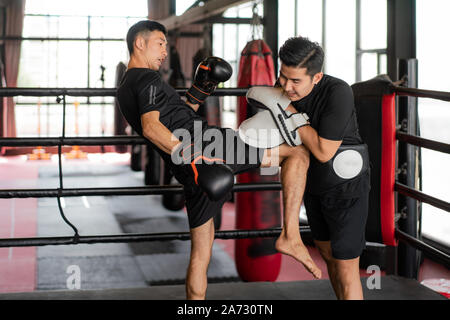 Giovani asiatici Kick boxer colpire con il ginocchio destro al trainer professionale nella boxe studuim in background in palestra per il fitness. Boxer stanno colpendo il sa Foto Stock