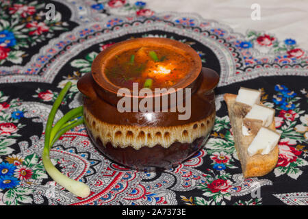 Borsch - zuppa di cavolo, barbabietole e carne di maiale - in una pentola di creta con lardo su un pezzo di pane e una piuma di cipolla verde Foto Stock