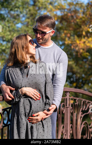 Felice coppia giovane con donna incinta in piedi insieme in un abbraccio come culle di lei il suo bambino bump nelle sue mani Foto Stock