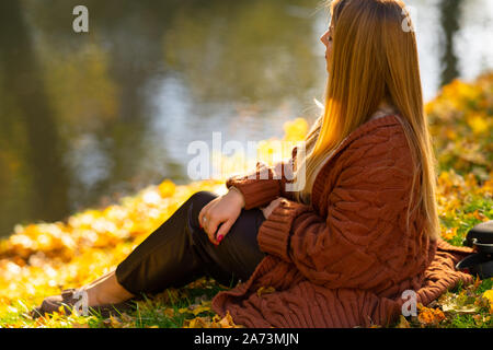 Moda giovane donna di relax all'aperto in un parco in autunno seduto sulla sponda di un fiume sul prato con sparsi in foglie di giallo Foto Stock