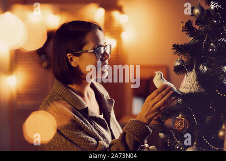 Giovane donna felice in occhiali in piedi vicino all'albero di Natale e guardando alla meravigliosa Colomba bianca su di esso Foto Stock