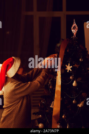 Giovane donna in Santa hat decorare albero di Natale con i giocattoli e preparazione per la vacanza a casa Foto Stock