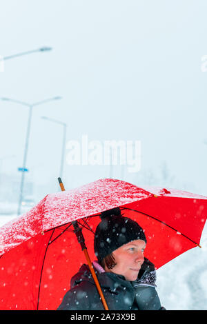 Piacevole donna parlando al telefono mobile sotto ombrellone rosso mentre in piedi in strada in inverno la neve Foto Stock