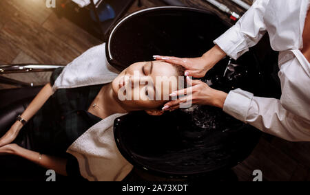 Stupendo carino giovane donna godendo di massaggio della testa mentre parrucchiere professionale di applicare lo shampoo capelli. Chiusura del parrucchiere mani capelli di lavaggio Foto Stock