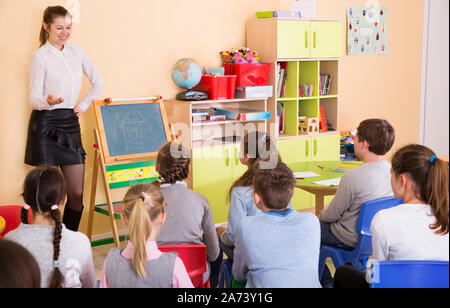 Giovani insegnanti di sesso femminile in piedi alla lavagna di classe, conducendo la lezione con i bambini Foto Stock