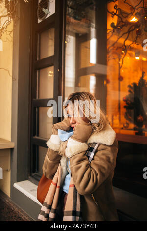 Bella ragazza bionda la sensazione di freddo, cercando di proteggere se stessa dal congelamento della città sul giorno d'inverno. Giovane donna caucasica attesa per amico sulla s Foto Stock