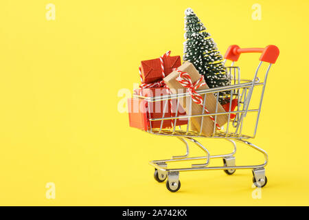 Carrello pieno di varie confezioni regalo e un albero di Natale su sfondo giallo. Natale il concetto di vendita. Foto Stock