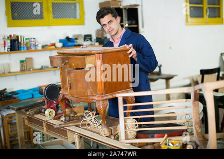 L'uomo carpenter indossando tute blu da ripristinare mobili in studio di restauro Foto Stock