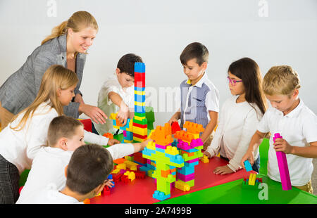 Gruppo di schoolkids giocando con mattoni giocattolo in cerchio con il loro allegro insegnante femmina Foto Stock