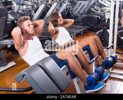 Ben addestrato uomo coach assistere gioiosa giovane donna per fare esercizio di abs sit ups in palestra indoor Foto Stock