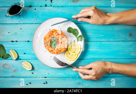 Deliziosa di avocado e salate Tartara di salmone su blu sullo sfondo di legno. Mani femminili tenere forchetta e coltello. Vista superiore Foto Stock