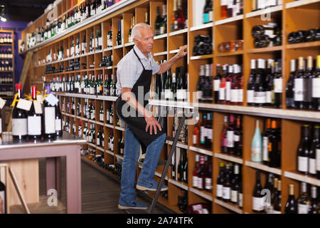 Anziani, maschi vignaiolo tenendo la bottiglia di vino mentre in piedi sulla scaletta vicino a ripiani in negozio Foto Stock