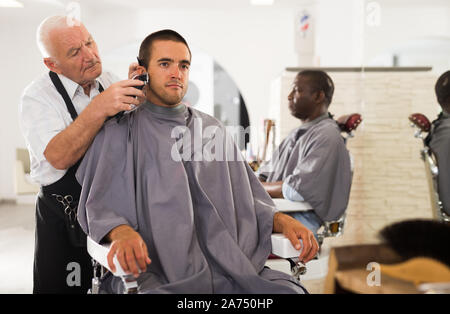 Giovane uomo getting haircut con clipper professionale da abili di età barbiere in salone Foto Stock