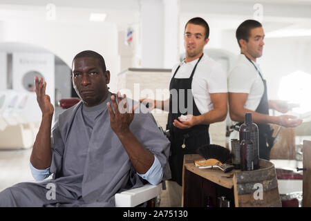 Ritratto di scioccato americano africano seduto nella sedia barbiere con confusi giovani parrucchiere dietro di lui Foto Stock