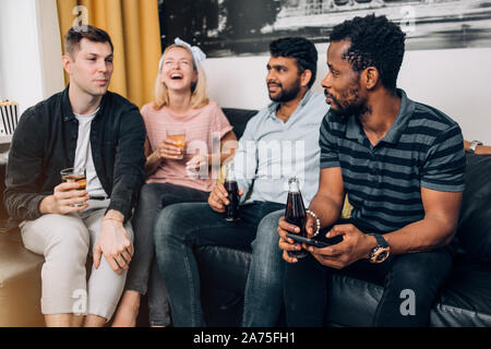 Gruppo di diverse gioiosa amici divertendosi a casa partito, allegramente a ridere, gustando un drink seduti sul divano nel soggiorno. Persone, Tempo libero e ven. Foto Stock