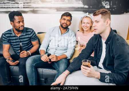 Gruppo di diverse gioiosa amici divertendosi a casa partito, allegramente a ridere, gustando un drink seduti sul divano nel soggiorno. Persone, Tempo libero e ven. Foto Stock