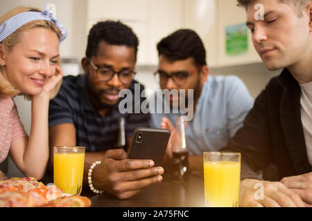 Quattro amici seduti al tavolo, di mangiare pizza e bere bevande analcoliche, la lettura di un messaggio di testo sullo smartphone, prove nuova applicazione telefono insieme. Mult Foto Stock