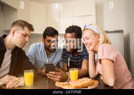 Quattro amici seduti al tavolo, di mangiare pizza e bere bevande analcoliche, la lettura di un messaggio di testo sullo smartphone, prove nuova applicazione telefono insieme. Mult Foto Stock