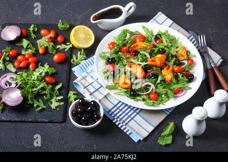 La rucola pinze di granchio insalata con olive nere, pomodorini e cipolle rosse servite su una piastra bianca, su una tavola di cemento con ingredienti, vista da un Foto Stock