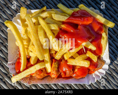 Delizioso piatto di cartone riempito con ketchup e patatine fritte rivestito accompagnato da currywurst, un tedesco specialità culinaria, mangiare all'aperto Foto Stock