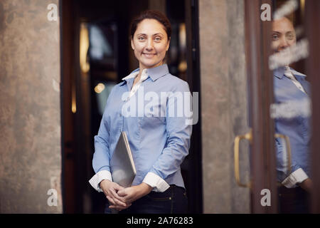 Ritratto di fiducioso imprenditrice matura in maglietta blu holding laptop nelle sue mani e sorridente in telecamera in ufficio Foto Stock