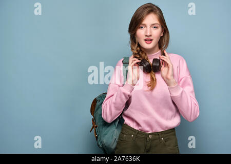 Una ragazza in un maglione rosa con uno zaino sorge su uno sfondo blu e trattiene le cuffie con le mani sul suo collo. Foto Stock