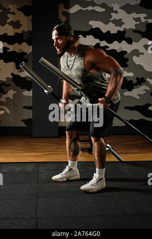 Uomo muscolare in uno sport uniforme e un cappello da baseball si erge nel profilo con barbells da barbell nelle sue mani Foto Stock