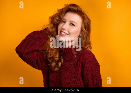 Redhead curly ragazza in un maglione di Borgogna si erge raddrizza i suoi capelli e sorrisi ampiamente contro uno sfondo giallo Foto Stock