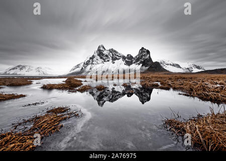 Coperto di neve e la formazione di montagna è riflessa in uno stagno, ciuffi di erba in e intorno allo stagno, lame di erba sono parzialmente spostata dal vento Foto Stock