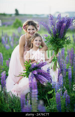 Madre e figlia la raccolta di fiori di lupino in un bellissimo campo sul tramonto. Bella ragazza in abito viola tenendo un lupino al tramonto sul campo. Il Foto Stock