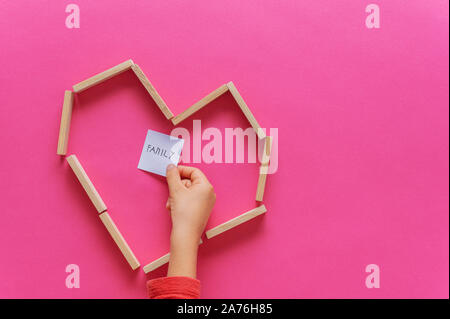 Forma di cuore fatta di pioli di legno con bambino mettendo mano post it carta con famiglia scritto su di esso all'interno del cuore. Su sfondo rosa. Foto Stock