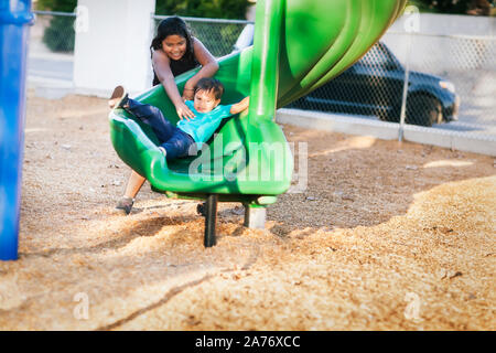 Vecchia sorella aiutare il suo fratello più giovane andare giù un twisted scorrere in un parco giochi per bambini. Foto Stock