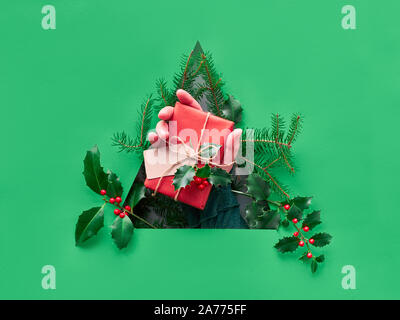 Tonica libro verde sullo sfondo di Natale con foro triangolare nel mezzo. Mano che tiene avvolto un regalo di Natale con la linguetta di cartone e holly ramoscelli Foto Stock