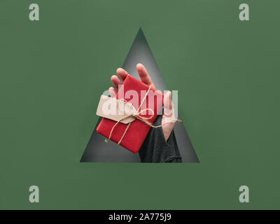 Il verde e il rosso della carta sullo sfondo di Natale con foro triangolare nel mezzo. Mano che tiene avvolto un regalo di Natale con la linguetta di cartone. Foto Stock