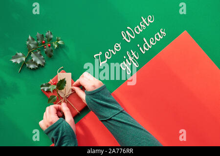 Creative appartamento panoramico giaceva sul verde e carta rossa con lunghe ombre . Femmina tenendo le mani avvolti confezione regalo decorate con agrifoglio. Eco-friendly Xmas, Foto Stock