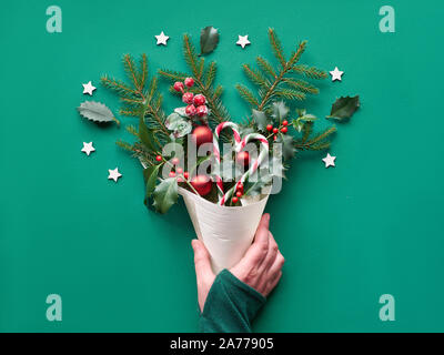 Creative piatto di Natale giaceva sul libro verde. Mano che tiene il cono di impiallacciatura naturale con decorazioni di Natale: abete e holly ramoscelli, baubles, candy canes, legno Foto Stock