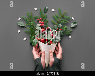 Creativi piatti di Natale i laici in verde e in rosso sulla carta grigia. Mano che tiene il cono di impiallacciatura naturale con decorazioni di Natale: abete e holly ramoscelli, baubles, cand Foto Stock