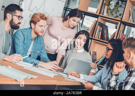 Startupers lavorando insieme in ufficio seduti a discutere di progetto mentre la donna incinta in piedi guardando il laptop con un collega Foto Stock