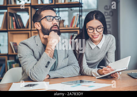 Startupers lavorando insieme a Office boss seduto pensieroso mentre segretario a prendere appunti sulle sue idee allegro Foto Stock