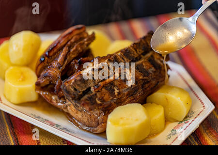 Cotto al forno costolette di maiale con patate giacente su di un piatto di festa con fiori e su una tabella con un etnica tovaglia striato, versata su un cucchiaio di salsa Foto Stock