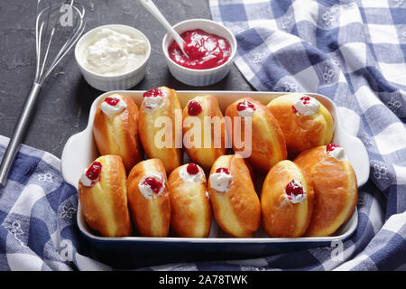 Close-up di Berliner Pfannkuchen, Tedesco Ciambelle con marmellata di lamponi e ripieni di crema in una teglia su una tabella di calcestruzzo con marmellata e crema in un arco Foto Stock