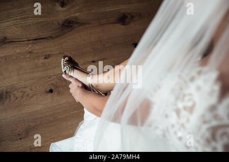 Elegante femmina mette le mani su scarpe matrimonio sullo sfondo un abito bianco, la mattina di nozze preparato in casa. Foto Stock