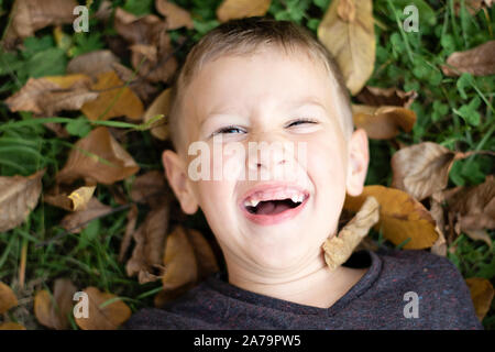 Ritratto di un giovane felice di pre-scuola ragazzo sdraiati sull'erba e foglie secche e ridere in parco pubblico in autunno. Ragazzo caucasico divertendosi in autunno. Foto Stock